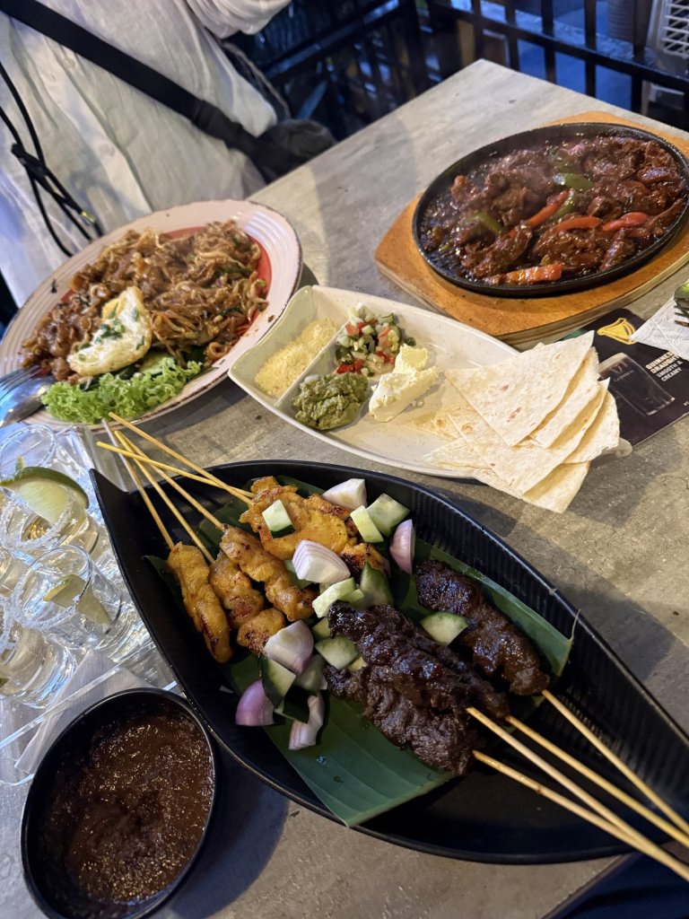 Satay-Spieße mit Erdnusssoße, Fladenbrot, Salat und Pfannengericht auf einem Tisch