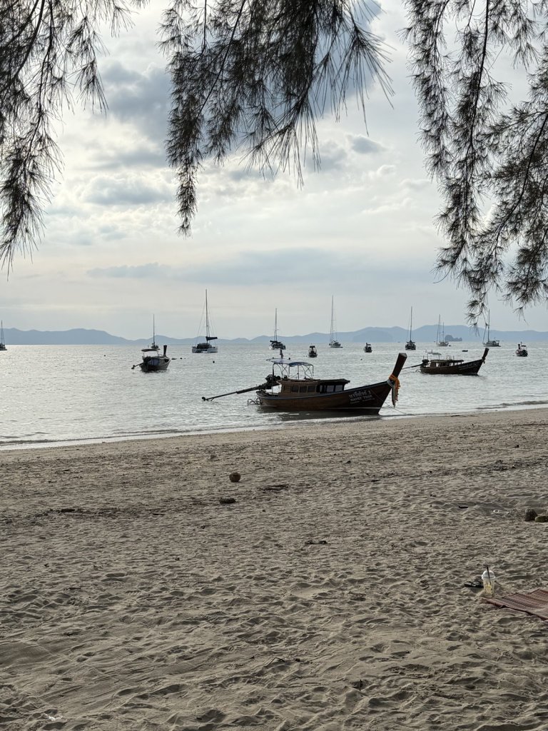 Longtail-Boot am Sandstrand, dahinter Inseln und Segelboote im Dunst