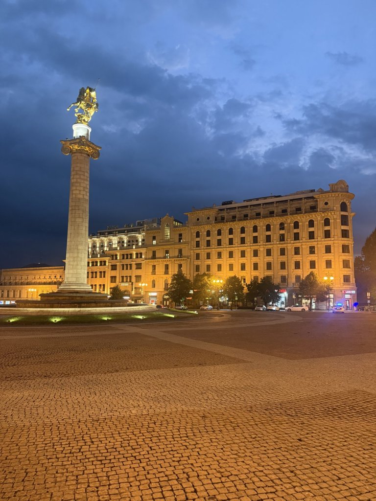 Freiheitsplatz in Tiflis mit goldener Statue auf einer Säule und beleuchteten Gebäuden unter dunklen Wolken