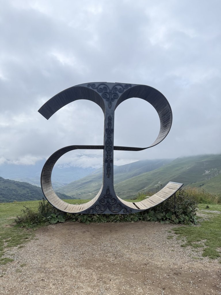 Metallskulptur des ossetischen Buchstabens Æ auf einer Wiese mit Blick ins Tal