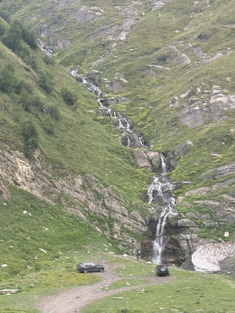 Wasserfall an einem grünen Berghang, unten parken zwei Autos