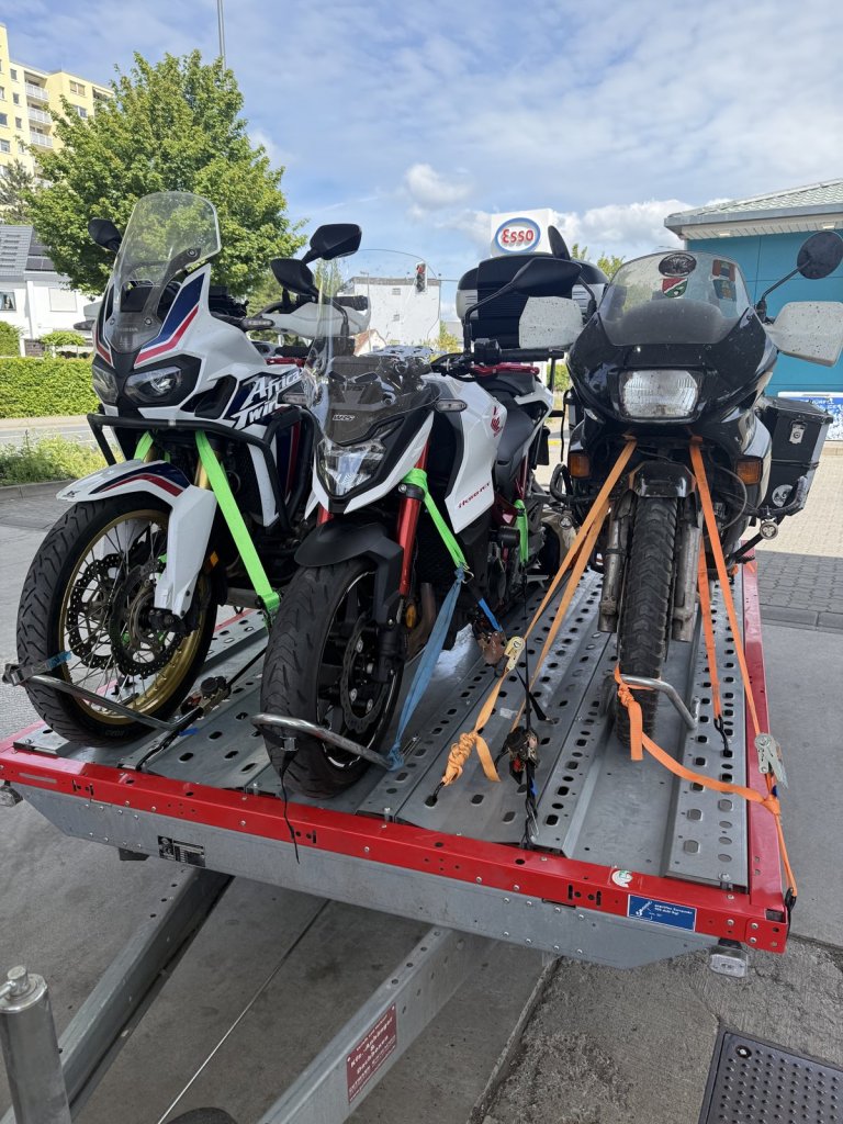 Drei Motorräder mit Spanngurten auf einem roten Anhänger an einer Tankstelle
