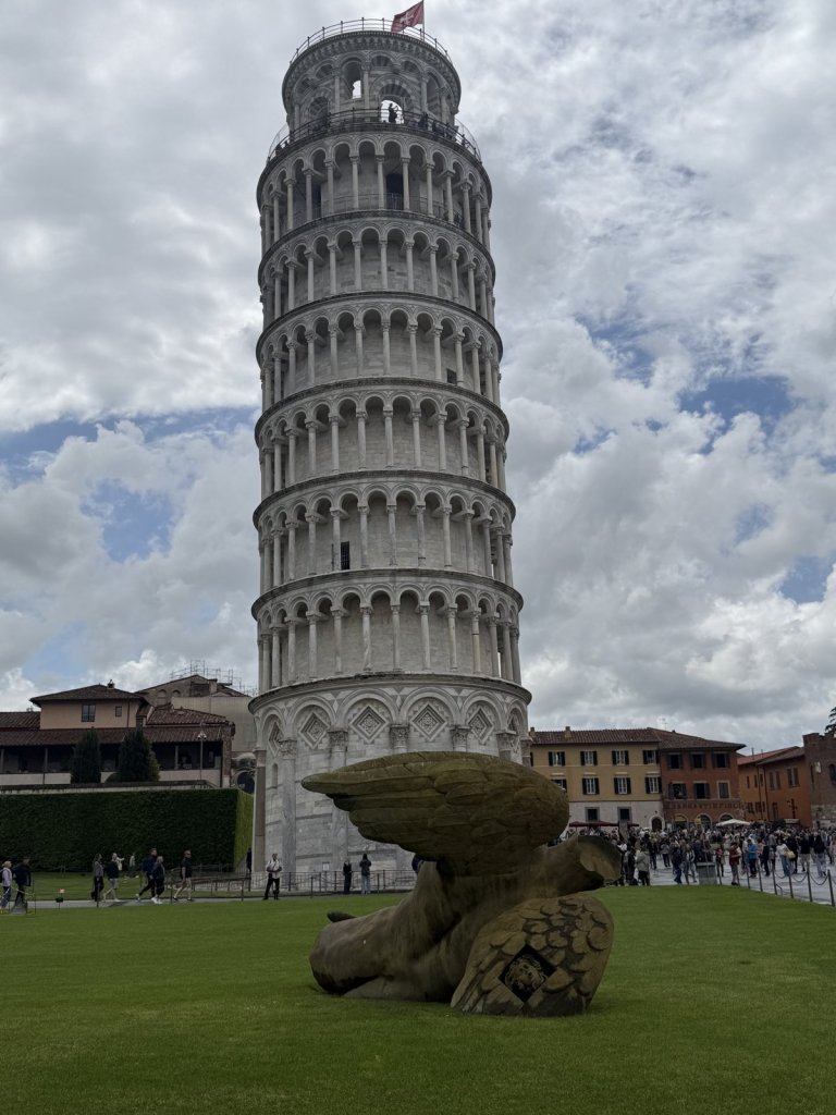 Schiefer Turm von Pisa über einer Wiese mit einer geflügelten Skulptur