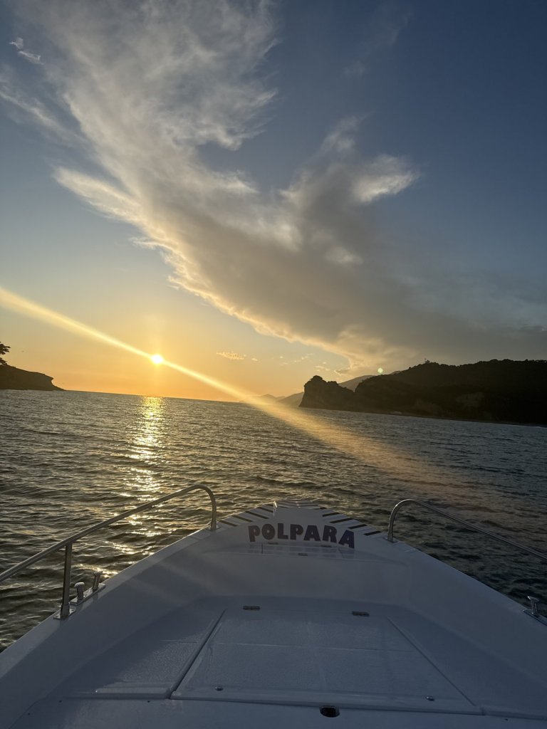 Sonne geht über dem Meer zwischen zwei Landzungen unter, im Vordergrund das Boot