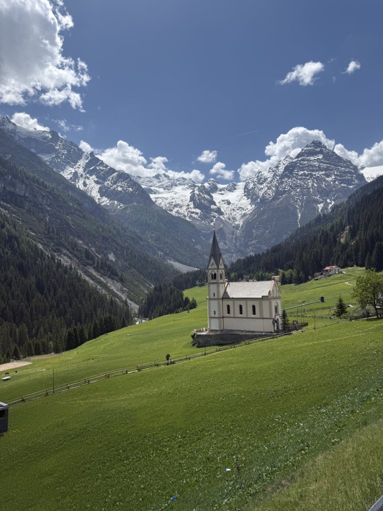 Weiße Kapelle auf einer grünen Wiese vor schneebedeckten Gipfeln