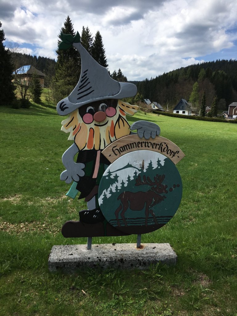 Holzschild mit Wichtel und Elch auf einer Wiese vor bewaldeten Hügeln