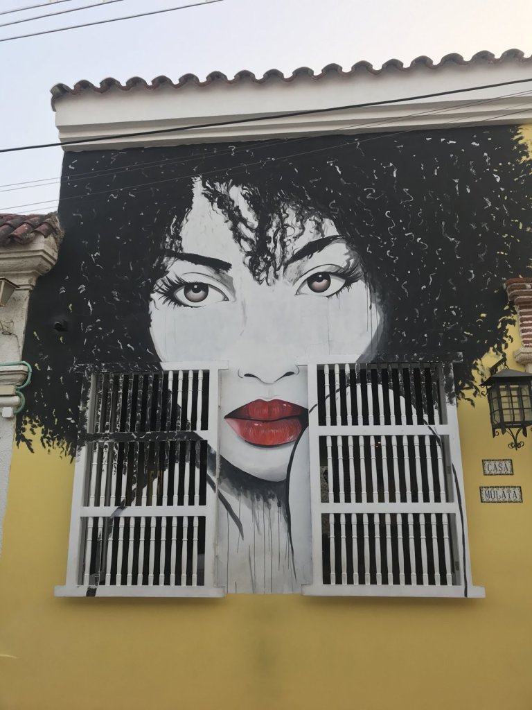 Wandbild einer Frau mit Locken und rotem Mund rund um ein vergittertes Fenster